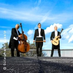 Drei Musiker in eleganten Anzügen, posieren mit Kontrabass, Gitarre und Cello vor blauem Himmel.
