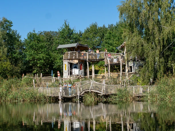 ArkaZien Erlebniswelt Region Leipzig - Seeterrassen mit Forschertürmen Über dem ruhigen See erhebt sich eine Holzterrasse mit Forschertürmen, umgeben von üppiger Vegetation und grünen Bäumen.