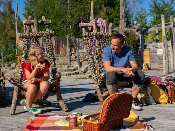Ojciec i córka cieszą się piknikiem na plecionych drewnianych krzesłach na słonecznym placu teatralnym, z koszem piknikowym i kolorowym kraciastym kocem przed sobą.