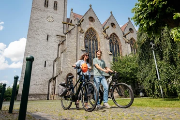 bad-laer-friedensroute-copyright-tol-foto-christoph-steinweg (6).jpg Zwei Menschen mit Fahrrädern vor einer historischen Kirche mit gotischen Fenstern.