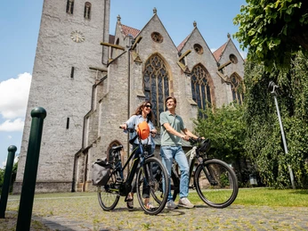 bad-laer-friedensroute-copyright-tol-foto-christoph-steinweg (6).jpg Zwei Menschen mit Fahrrädern vor einer historischen Kirche mit gotischen Fenstern.