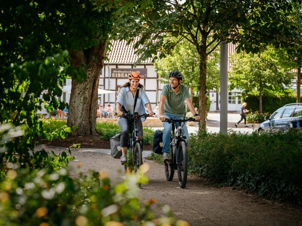 Radfahren in Bad Laer Zwei Radfahrende unterwegs auf einem schattigen Weg durch Bad Laer, umgeben von Grünflächen und Ortsbebauung.