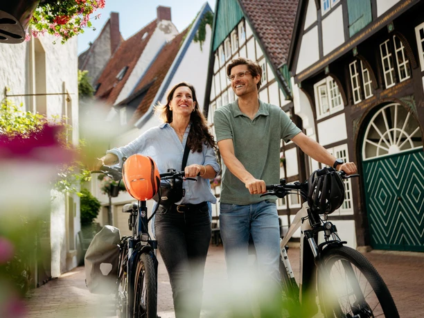 osnabrueck-friedensroute-altstadt-c-tol-foto-christoph-steinweg (1).jpg Paar radelt lächelnd durch eine von Fachwerkhäusern gesäumte kopfsteingepflasterte Straße.