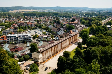 Gradierwerk in einer grünen Parklandschaft, umgeben von Stadthäusern und Hügeln im Hintergrund.