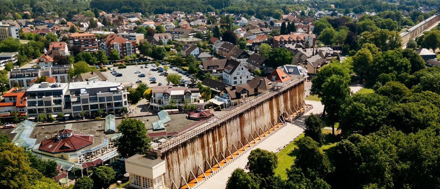 Gradierwerk in einer grünen Parklandschaft, umgeben von Stadthäusern und Hügeln im Hintergrund.
