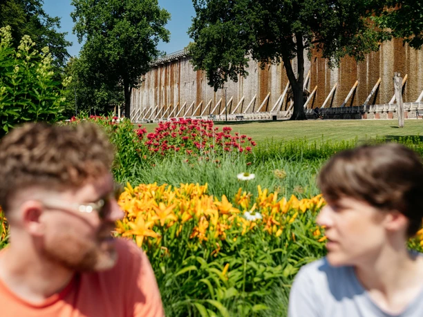 bad-rothenfelde-grenzgaengerroute-copyright-tol-foto-christoph-steinweg (3).jpg Zwei Personen vor einem alten Gradierwerk mit blühendem Garten im Vordergrund.