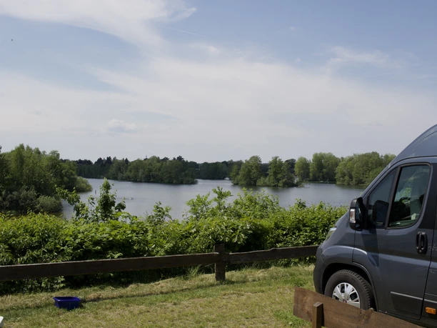 Campingplatz Badeinsel Ernst und Christa Pfaff Ein Campingplatz an einem See mit Bäumen entlang des Ufers. Ein grauer Van steht am Rand.