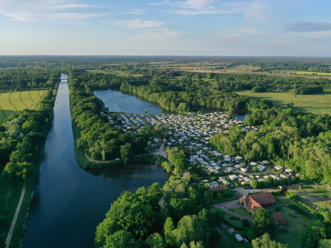 Campingplatz Forellensee Ein weitläufiger Campingplatz mit Wohnmobilen an einem großen See inmitten von grüner Natur.