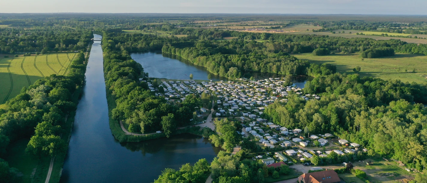 Campingplatz Forellensee Ein weitläufiger Campingplatz mit Wohnmobilen an einem großen See inmitten von grüner Natur.