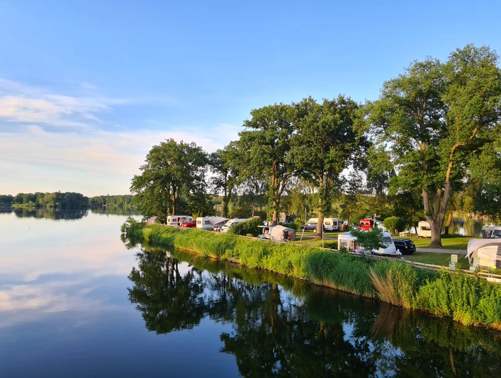 Camping Lanzer See_Halbinsel Das Bild zeigt eine Halbinsel am Lanzer See, gesäumt von Bäumen und Wohnmobilen. Die ruhige Wasseroberfläche spiegelt die Landschaft wieder.