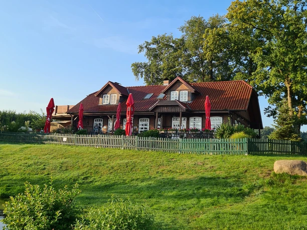 Camping Lanzer See_Gastro Eine rote Ziegeldach-Gaststätte mit roten Sonnenschirmen auf einer grünen Wiese am Lanzer See.