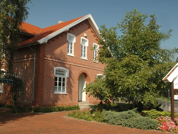 Landwirtschaftsmuseum Rhede (Ems) ©Gemeinde Rhede (Ems) .JPG Backsteingebäude mit rotem Dach, umgeben von Bäumen, Blumenbeeten und dem Schild des Landwirtschaftsmuseums.