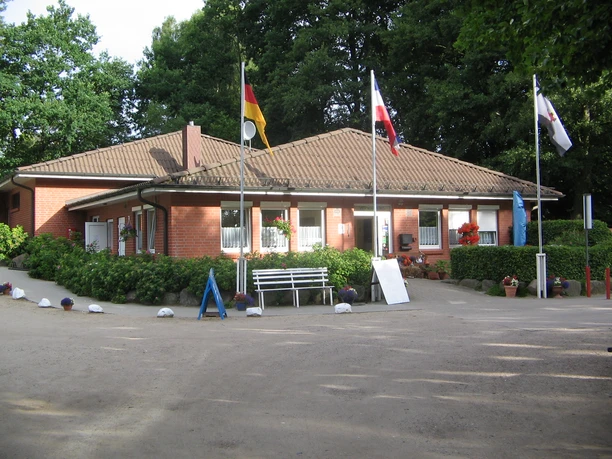 Campingplatz am Gudower See_Empfang Ein einstöckiges Backsteingebäude mit Dachziegeln, Flaggensäulen, umgeben von Bäumen.