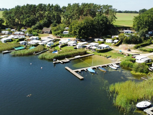 Campingplatz Groß Zecher_von oben Campingplatz mit Wohnwagen und Zelten am See mit Bootsanlegern, umgeben von Bäumen und Grünflächen.