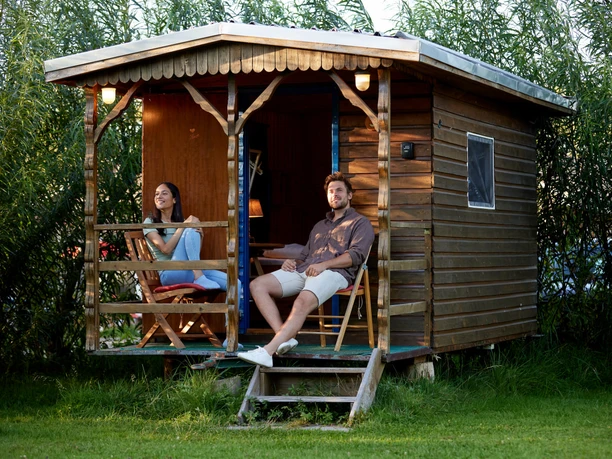 Wakenitz-Camp Eine Frau und ein Mann sitzen entspannt auf der Veranda einer kleinen Holzhütte im Grünen.