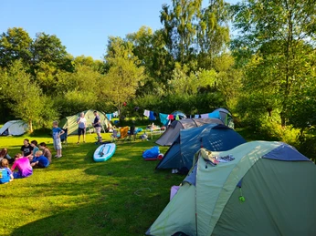 Wakenitz-Camp_Zeltplatz Zelte stehen auf grüner Wiese, Menschen machen Picknick, Wälder im Hintergrund, sonniges Wetter.