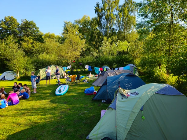 Wakenitz-Camp_Zeltplatz Zelte stehen auf grüner Wiese, Menschen machen Picknick, Wälder im Hintergrund, sonniges Wetter.
