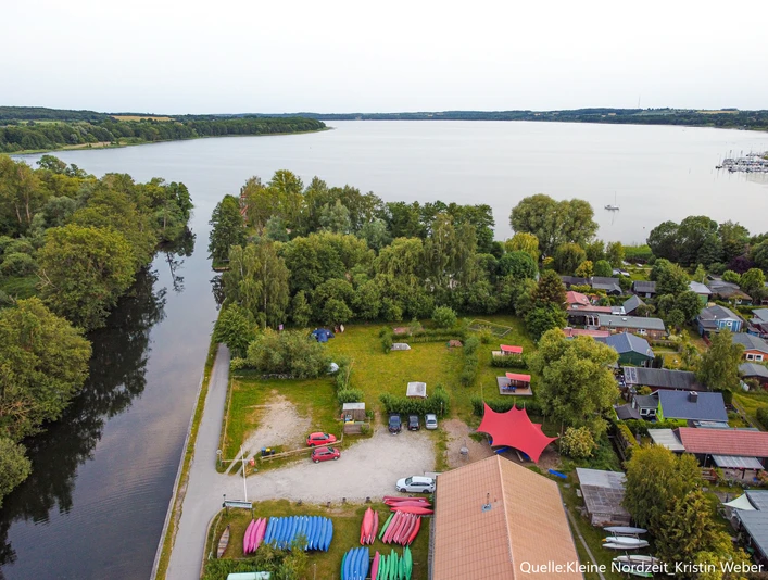 Wakenitz-Camp Blick auf ein Gewässer, umliegende grüne Vegetation, Boote am Ufer und ein Campingplatz.
