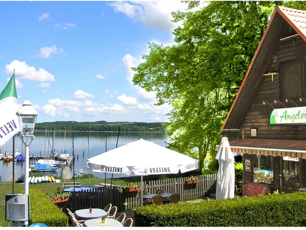 Naturcamping Buchholz_Mühle Ein Campingplatz am See mit grünem Ufer, Segelbooten und einem holzverkleideten Restaurant im Vordergrund.