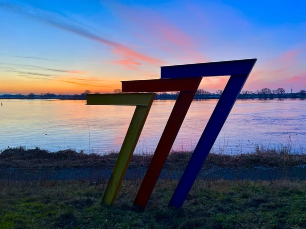 Wohnmobilstellplatz Geesthacht_777 Farbenfrohe Skulptur in Form der Zahl "777" am Ufer, im Hintergrund ein ruhiger Sonnenuntergang.