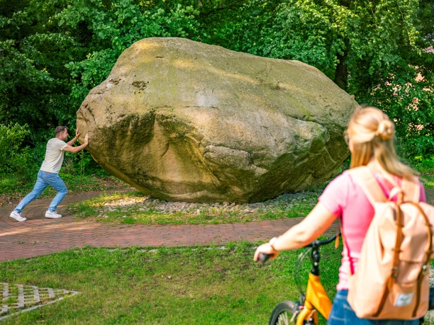 Koloss von Hüven – Radfahren Hümmling ©Emsland Tourismus GmbH (2).jpg Ein Mann stemmt sich gegen einen großen Findling, während eine Radfahrerin das Geschehen beobachtet.