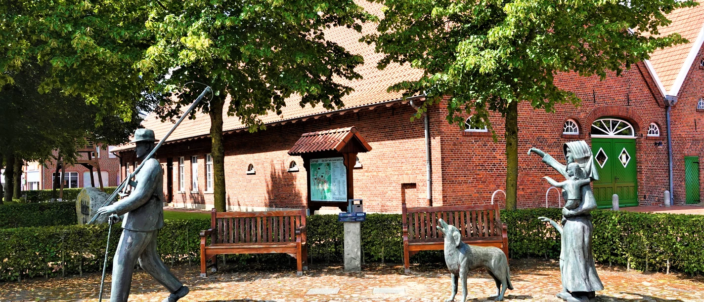 Krulls Hus in Lorup: Wo heute das gut 100 Jahre alte Heimathaus steht, gibt es seit mindestens 1000 Jahren eine Hofstelle, die zwischen 1749 und 1982 der Familie Krull gehörte – ein guter Ort also, um die Dorf-Geschichte zu bewahren. Backsteingebäude mit grünem Tor, davor Bronzefiguren einer Familie mit Hund unter schattigen Bäumen.