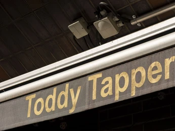Toddy Tapper Ein Schild mit der Aufschrift 'Toddy Tapper' in goldener Schrift vor einer dunklen Hintergrundstruktur.A sign with the inscription 'Toddy Tapper' in gold lettering against a dark background structure.