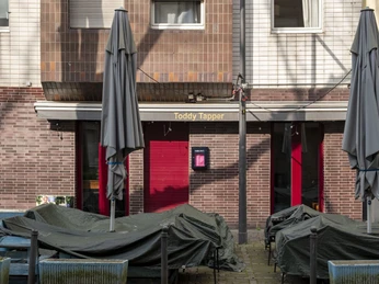 Toddy Tapper Vor einem Backsteingebäude sind Terrasse und Möbel mit Abdeckungen versehen. Zwei graue Sonnenschirme sind geschlossen.In front of a brick building, the terrace and furniture are covered. Two gray sunshades are closed.