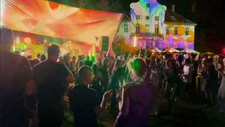 parkbeben-kulturgut -poggenhagen Personen tanzen bei Nacht vor einer beleuchteten Bühne, im Hintergrund ein farbig angestrahltes Gebäude.