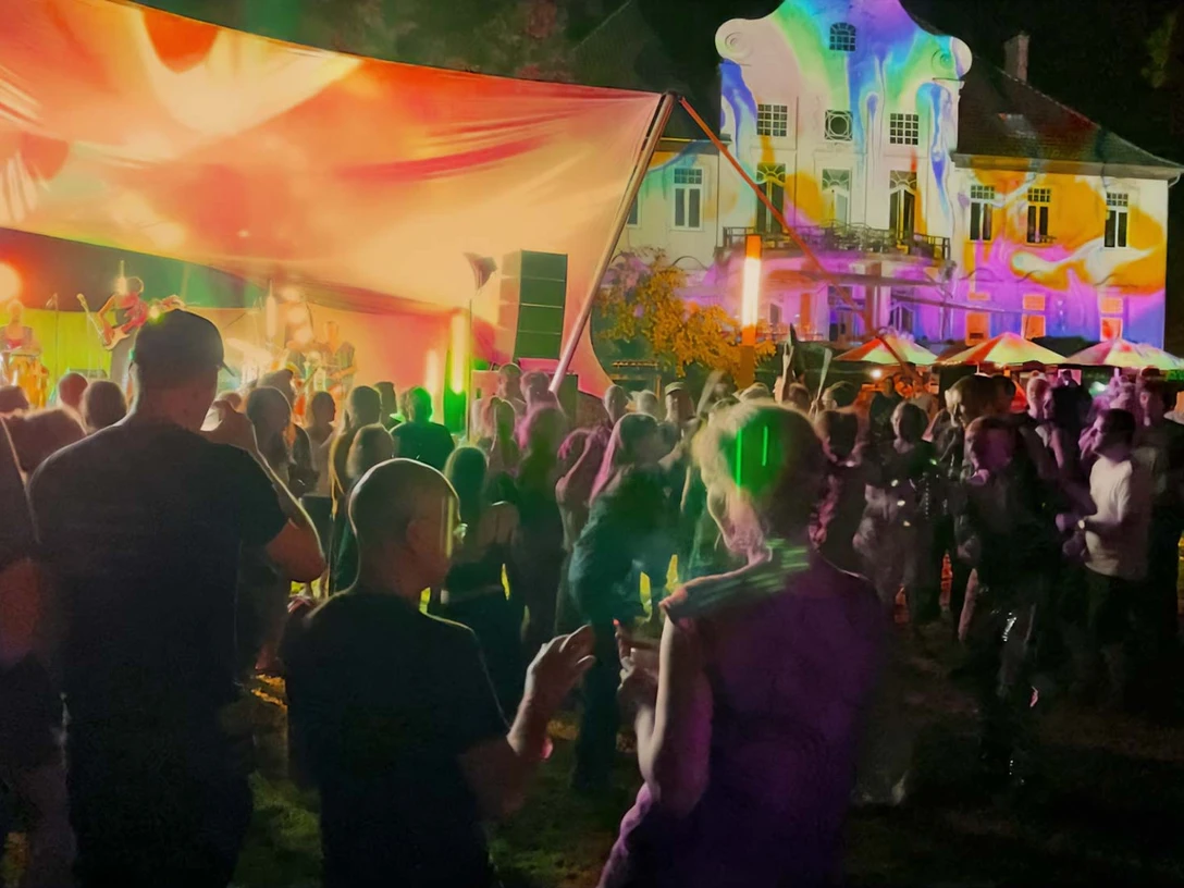 parkbeben-kulturgut -poggenhagen Personen tanzen bei Nacht vor einer beleuchteten Bühne, im Hintergrund ein farbig angestrahltes Gebäude.