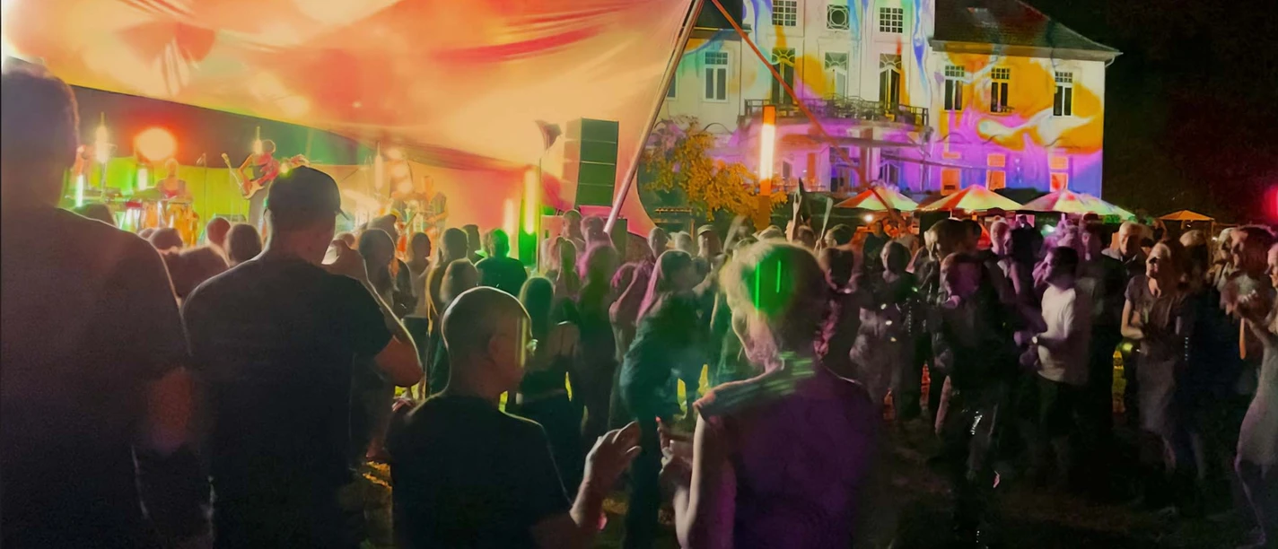 parkbeben-kulturgut -poggenhagen Personen tanzen bei Nacht vor einer beleuchteten Bühne, im Hintergrund ein farbig angestrahltes Gebäude.