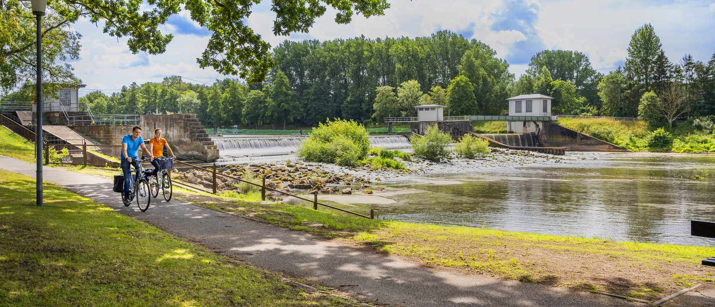 Wasserfall Hanekenfähr, Lingen (Ems) – Radfahren im Emsland ©Emsland Tourismus GmbH.jpg Zwei Radfahrende auf Weg am Wasserfall Hanekenfähr bei Lingen, umgeben von grüner Flusslandschaft.
