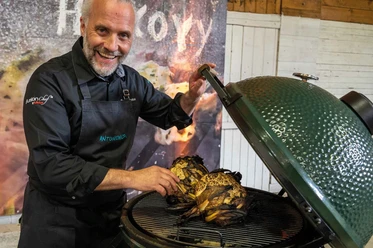 FIRE&FOOD BBQ WEEK Ein freundlicher Koch präsentiert stolz zwei gegrillte Kohlköpfe auf einem großen, grünen Grill.