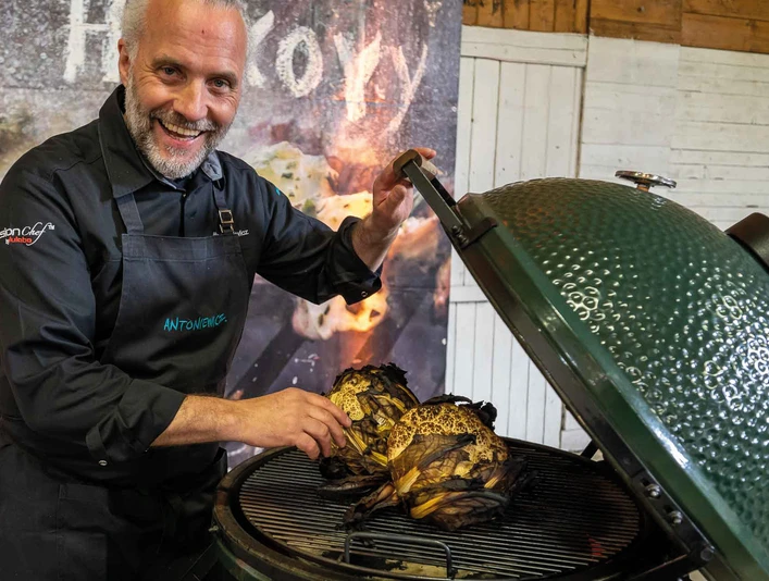 FIRE&FOOD BBQ WEEK A friendly chef proudly presents two grilled cabbages on a large green grill.