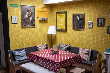 The Bär Das Bild zeigt einen Raum mit gelben Tapeten, einer Sitzecke mit Polsterkissen und dekorativen Bildern an der Wand. Ein Tisch mit rotem Punktetuch steht zentral, ergänzt durch bequeme Holzstühle.