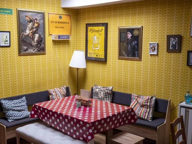 The Bär Das Bild zeigt einen Raum mit gelben Tapeten, einer Sitzecke mit Polsterkissen und dekorativen Bildern an der Wand. Ein Tisch mit rotem Punktetuch steht zentral, ergänzt durch bequeme Holzstühle.