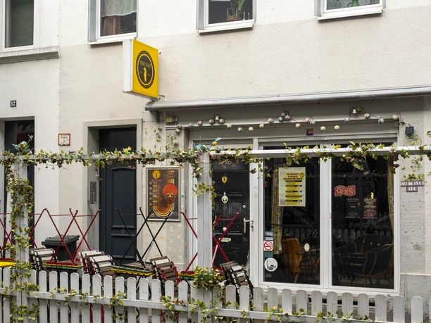 The Bär Ein Café in Köln mit weißer Fassade und einladendem Außenbereich. Bunte Stühle stehen vor einem weißen Zaun, der mit grünen Pflanzen dekoriert ist.