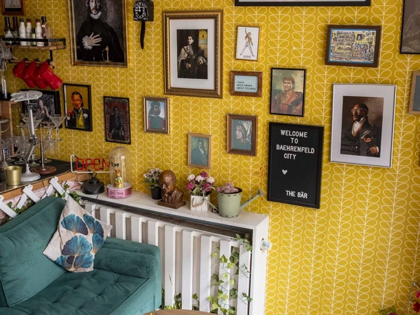 The Bär Numerous works of art hang in a room with yellow patterned wallpaper. A petrol-colored sofa, adorned with stylish cushions, stands in front of a narrow wall table. Plants and curiosities are placed on it. A small wooden table with books and magazines completes the artistic ensemble.