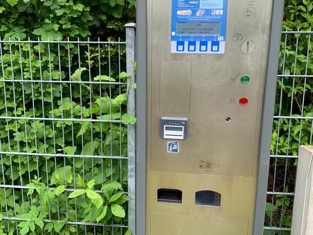 Wohnmobilstellplatz Mölln_Automat Ein Parkautomat vor einem grünen Zaun auf bewachsener Wiese. Der Automat ist silberfarben.