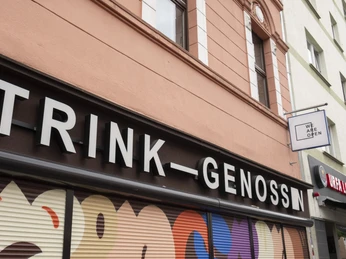 TRINK-GENOSSIN Der Schriftzug 'TRINK-GENOSSIN' auf einem Gebäude mit ansprechender moderner Schaufenstergestaltung.The lettering 'TRINK-GENOSSIN' on a building with an appealing modern shop window design.