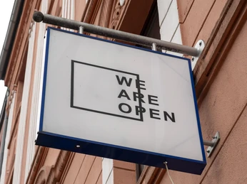 TRINK-GENOSSIN Ein rechteckiges Schild mit der Aufschrift „WE ARE OPEN“ hängt an einer rosafarbenen Hausfassade.A rectangular sign with the inscription "WE ARE OPEN" hangs on a pink-colored house facade.