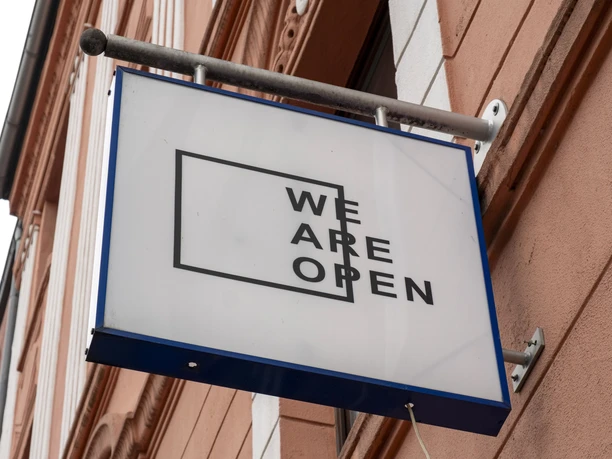 TRINK-GENOSSIN Ein rechteckiges Schild mit der Aufschrift „WE ARE OPEN“ hängt an einer rosafarbenen Hausfassade.