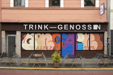 TRINK-GENOSSIN Fassade eines Kölner Lokals mit buntem Wandkunstwerk und leeren Tischen im Vordergrund.