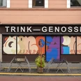 TRINK-GENOSSIN Fassade eines Kölner Lokals mit buntem Wandkunstwerk und leeren Tischen im Vordergrund.Facade of a Cologne restaurant with colorful wall art and empty tables in the foreground.