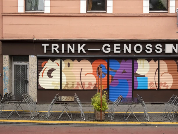 TRINK-GENOSSIN Fassade eines Kölner Lokals mit buntem Wandkunstwerk und leeren Tischen im Vordergrund.
