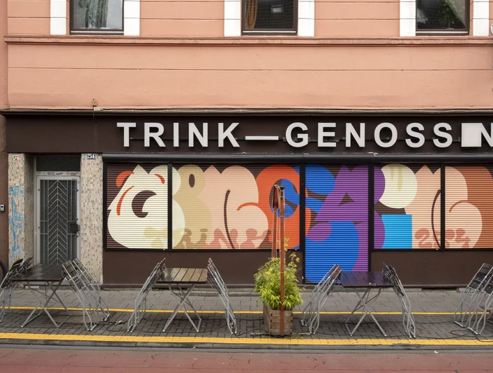 TRINK-GENOSSIN Fassade eines Kölner Lokals mit buntem Wandkunstwerk und leeren Tischen im Vordergrund.Facade of a Cologne restaurant with colorful wall art and empty tables in the foreground.