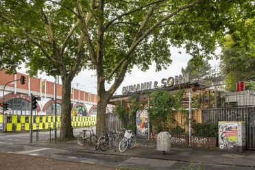 Bumann & Sohn Ein Außenbereich eines Veranstaltungsortes mit dem Namen "Bumann & Sohn" unter schattigen Bäumen, Fahrräder stehen geparkt.