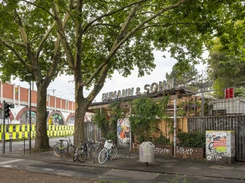 Bumann & Sohn Ein Außenbereich eines Veranstaltungsortes mit dem Namen "Bumann & Sohn" unter schattigen Bäumen, Fahrräder stehen geparkt.An outdoor area of an event venue called "Bumann & Sohn" under shady trees, bicycles parked.