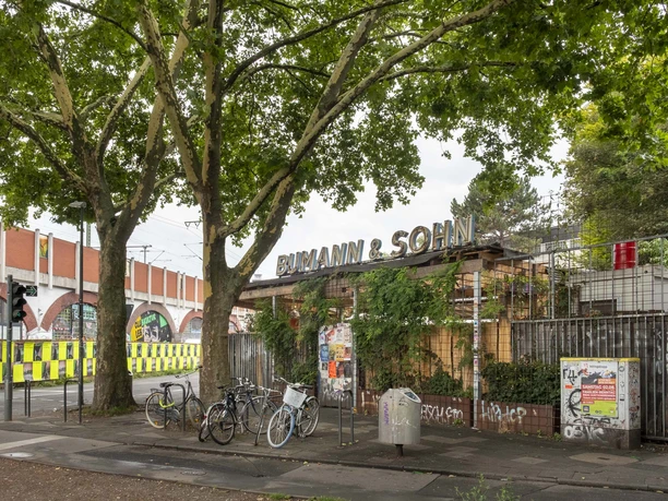 Bumann & Sohn Ein Außenbereich eines Veranstaltungsortes mit dem Namen "Bumann & Sohn" unter schattigen Bäumen, Fahrräder stehen geparkt.