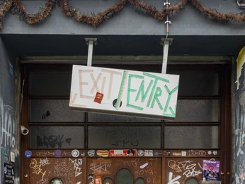 Bumann & Sohn Der Eingang zu einer Bar ist mit Grafitti und einem Schild "EXIT/ENTRY" dekoriert. Die Tür ist aus Holz, von Stickern und Kritzeleien überzogen, ein Beispiel kreativer Street Art.The entrance to a bar is decorated with graffiti and an "EXIT/ENTRY" sign. The door is made of wood, covered in stickers and doodles, an example of creative street art.